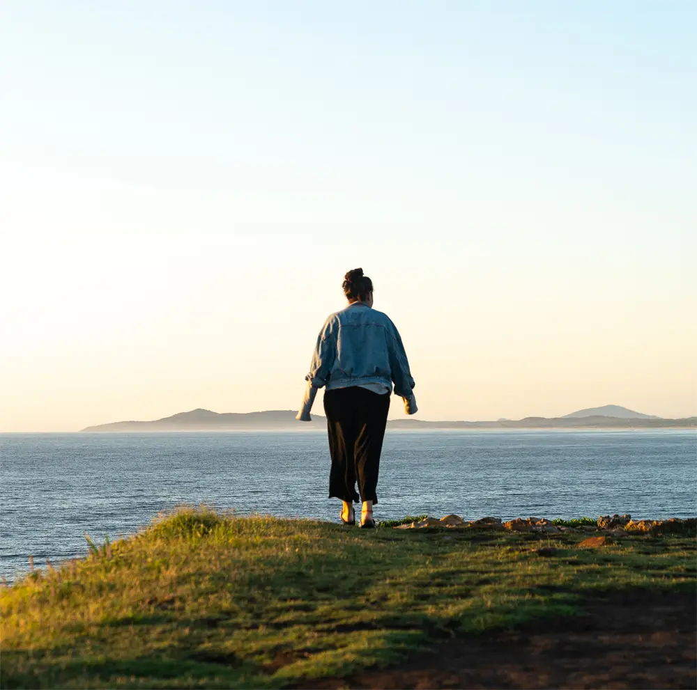 Woman in a denim jacket looking out at the ocean from a grassy cliff at sunset
